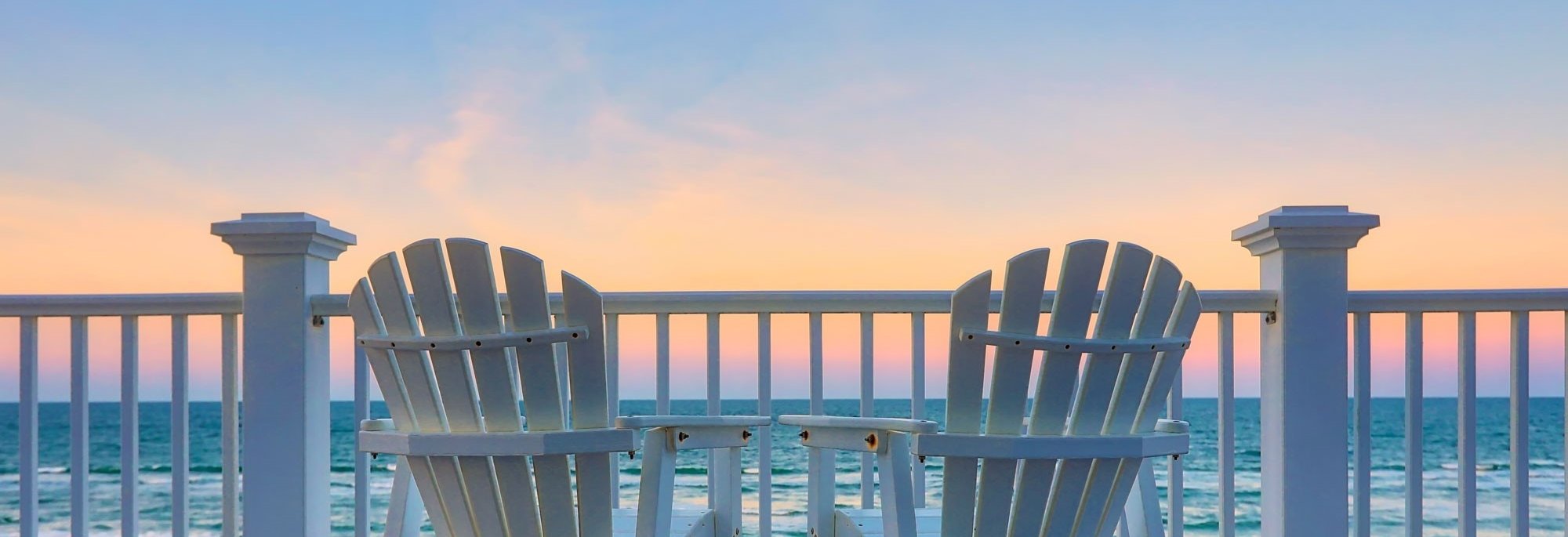 view of calm beach at sunset from behind Adirondack chairs and deck rails 